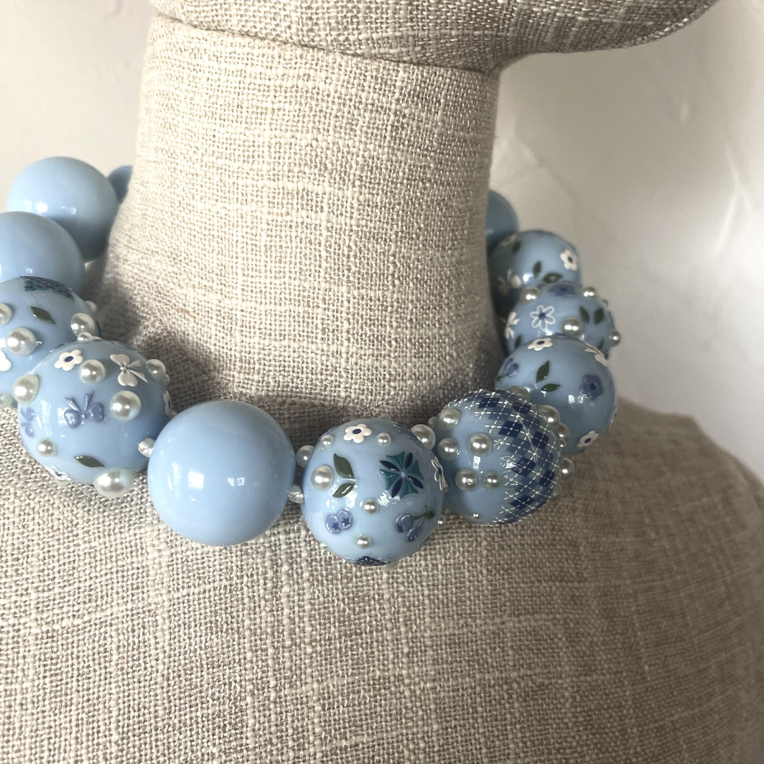 Baby‑blue acrylic bead necklace with floral and bow decorations displayed on a black jewelry bust.